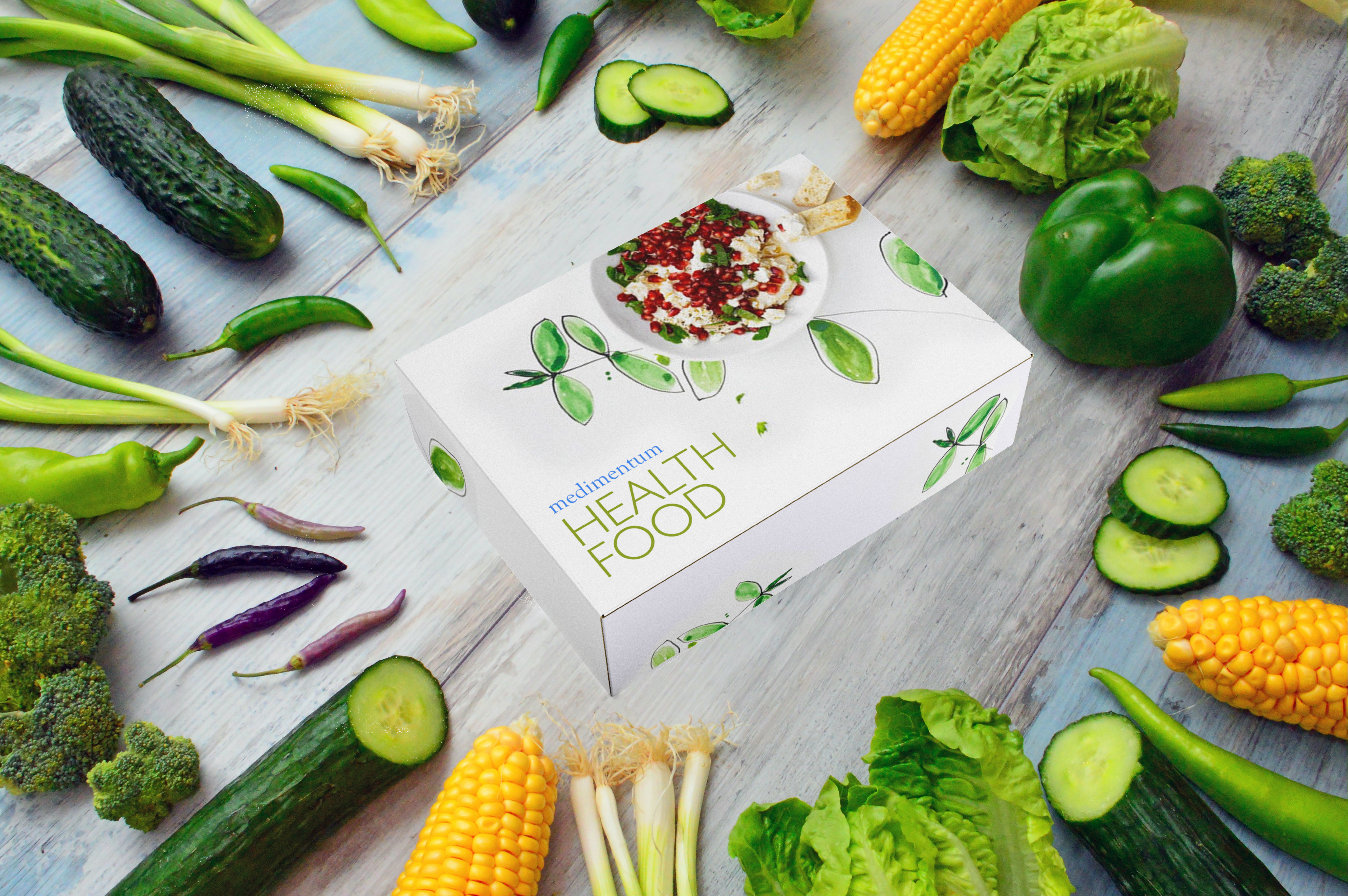 Food box from medimentumHealthFood.store between green vegetables on a wooden background.